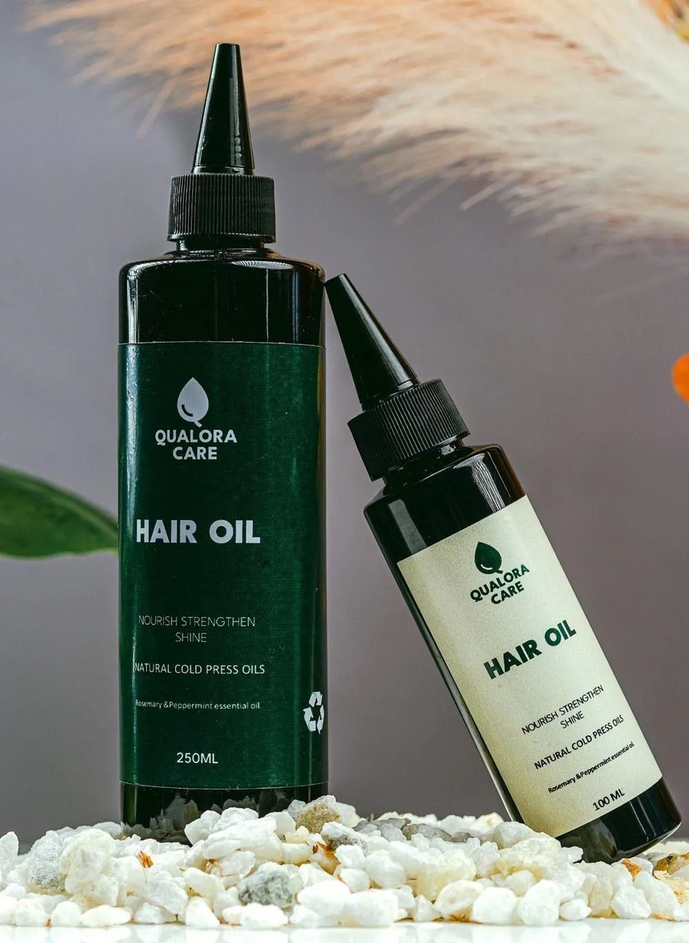 Qualora Care Hair Oil in 250 ml and 100 ml bottles on white stones, with pampas grass behind
