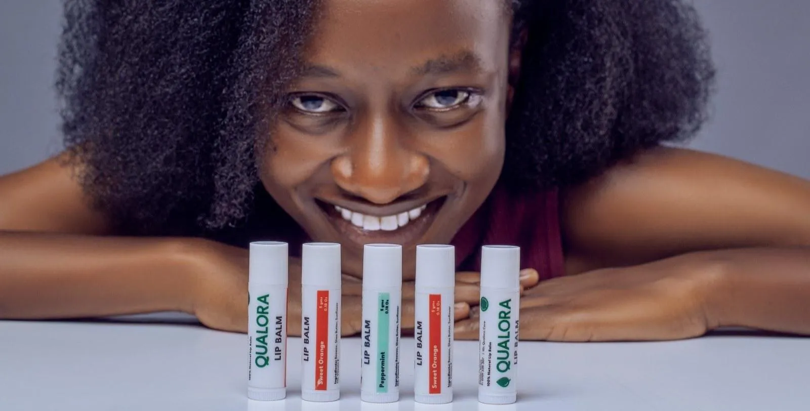 A smiling woman with natural hair rests her chin on her hands behind five Qualora Care lip balms